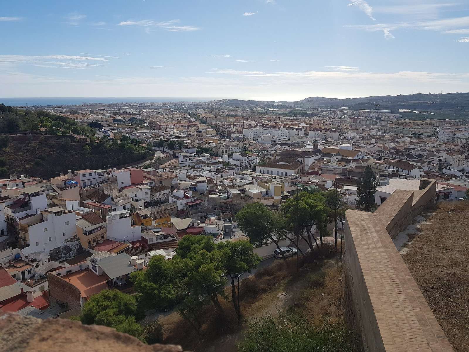 Vista panorámica de Vélez-Málaga desde una muralla, con casas blancas, el mar al fondo y montañas en el horizonte.