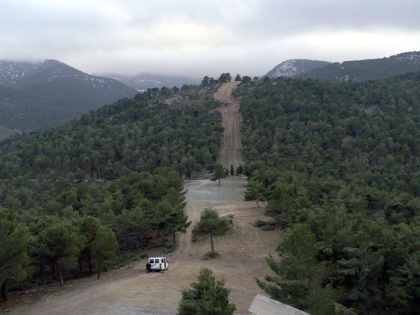 Pista de tierra entre pinares en la Sierra de Baza, con una furgoneta blanca y montañas con nieve y nubes al fondo.